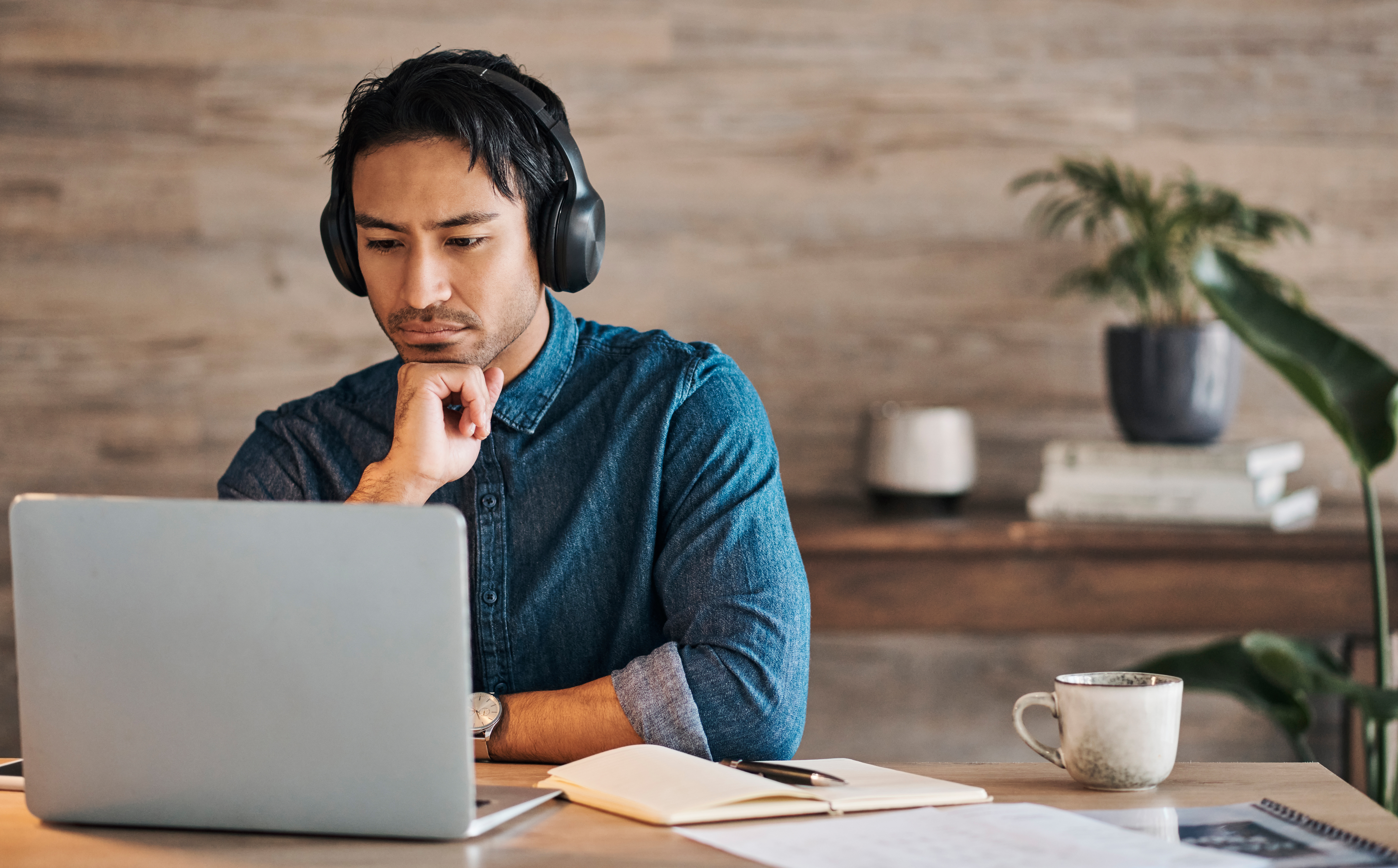 Businessman, headphones and laptop webinar in office with coffee on table, video call or watching video. Zoom call, video conference and male from Canada in online meeting with book and pen on desk.